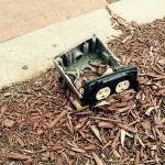 This outdoor receptacle has been destroyed. It is located in a planter bed next to a stall in a parking lot. It has been knocked over and completely obliterated. It should have been mounted on a 4x4 post  set in concrete.